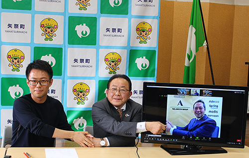 テーブル上で握手するボノ株式会社取締役 谷津氏と矢祭町 佐川町長、モニターに映る株式会社VSN代表取締役社長 川崎。背景には矢祭町のロゴ入りパネルと町旗。