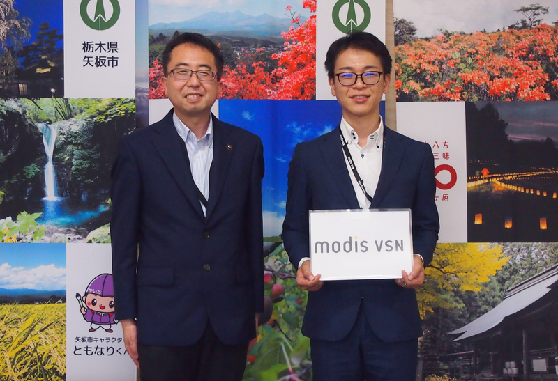 並んで立つ矢板市の齋藤淳一郎市長とModis VSN社員の秋本一樹。背景には矢板市の観光地や自然を紹介するパネルが並び、秋本が「modis VSN」と書かれたボードを持っている。