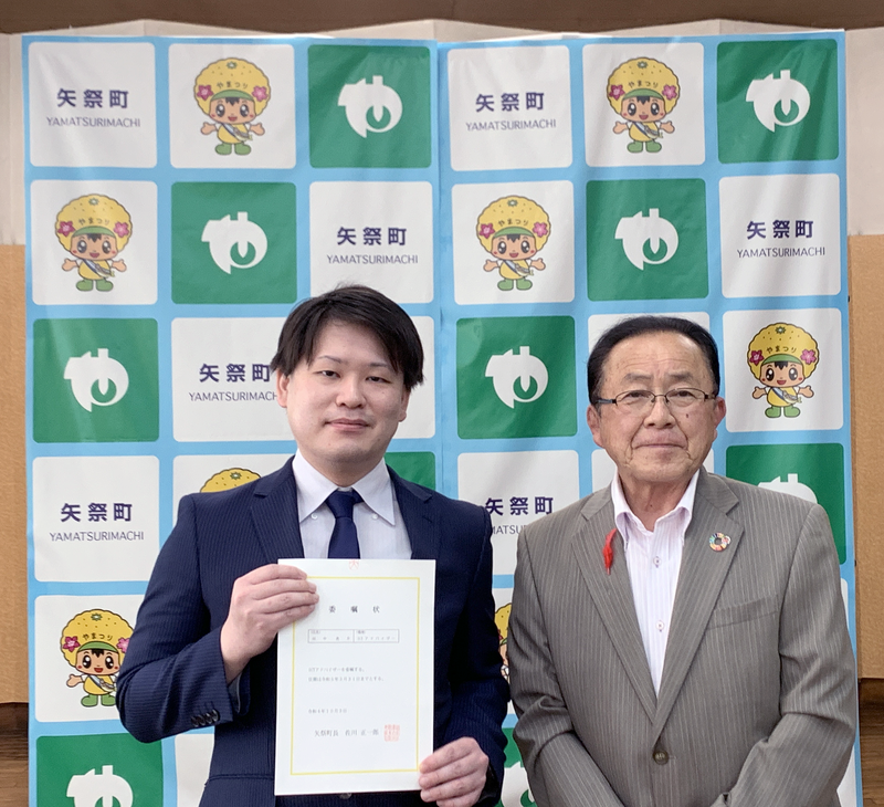 並んで立つModis社員の田中と矢祭町の佐川町長。田中は委任状を両手で持っている。背景には矢祭町のロゴ入りパネル。