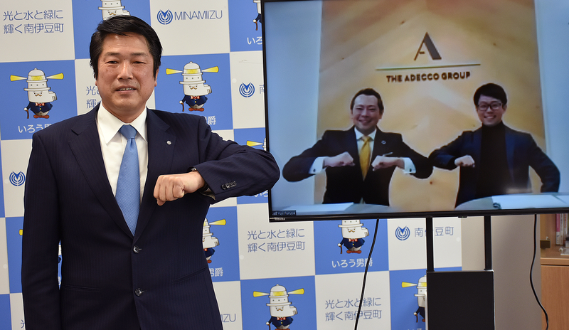 南伊豆町の岡部町長と、モニターに映るModis社長の川崎と株式会社ボノの谷津氏。背景には南伊豆町のロゴと「光と水と緑に輝く南伊豆町」の文字、ゆるきゃら「いろう男爵」の3つが書かれたパネルがかかっている。