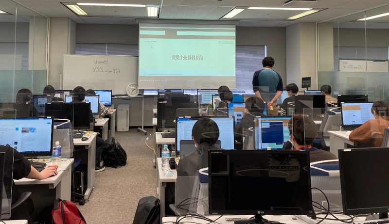 パソコンが並ぶ教室で複数の受講者が各々のモニターへ向かい作業している様子。前方のスクリーンには「競技開始」と表示されている。