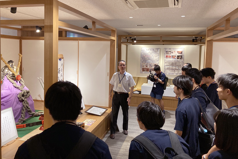 展示室でガイドによる説明を、複数の来場者が聞いている。