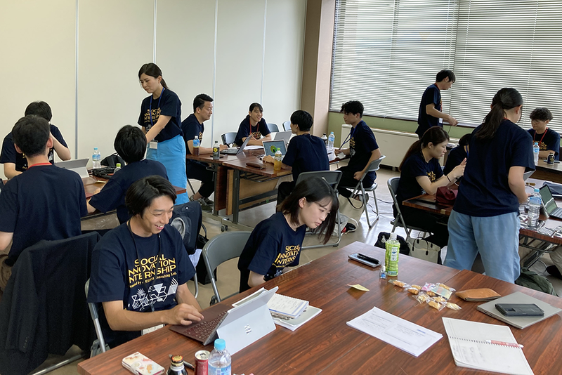 複数のグループがテーブルを囲み、ノートパソコンや資料を使ってグループワークをしている。