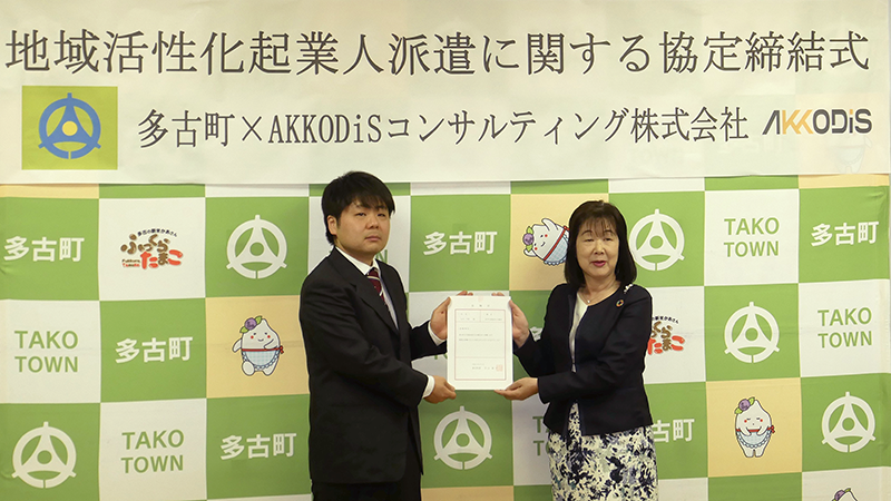 並んで立つ男性と女性。二人で一枚の書類を持っている。背後には「地域活性化企業人派遣に関する協定締結式 多古町×Akkodisコンサルティング株式会社」と書かれた帯が掛けられている。