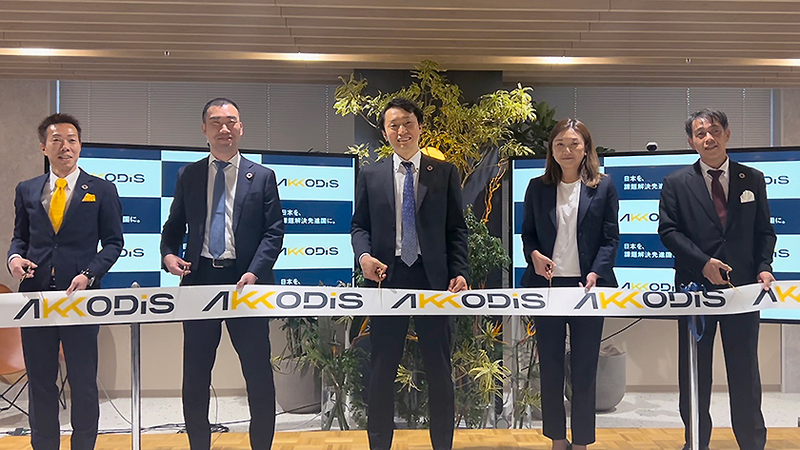 オフィス内で行われているテープカットセレモニー。背景には『AKKODIS』のロゴが表示された複数のモニターと観葉植物がある。5名の参加者が横一列に並び、ブランド名入りのテープを持っている。
