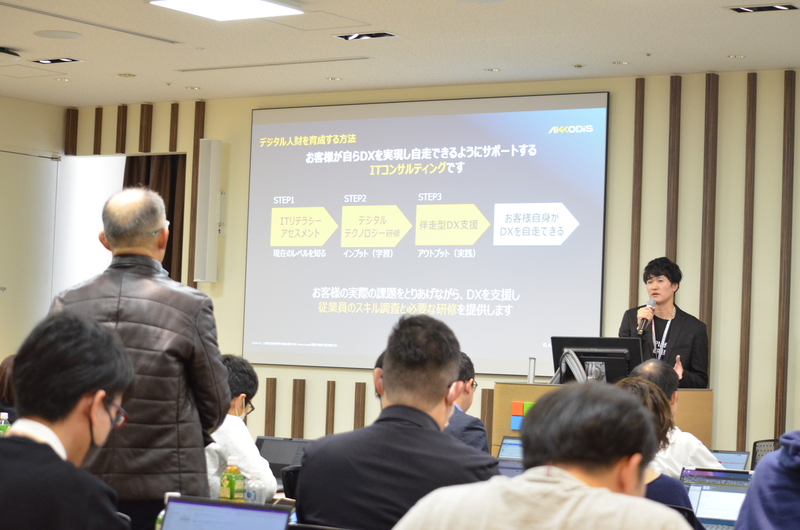 会議室でデジタル人材を育成する方法についてのプレゼンテーションの様子。登壇者はマイクを持ち説明し、参加者はノートパソコンを使ってメモを取っている。