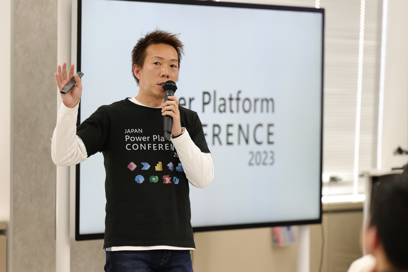 JAPAN Power Platform CONFERENCEで、男性の登壇者がマイクを持ち、片手を挙げながらプレゼンテーションを行っている。