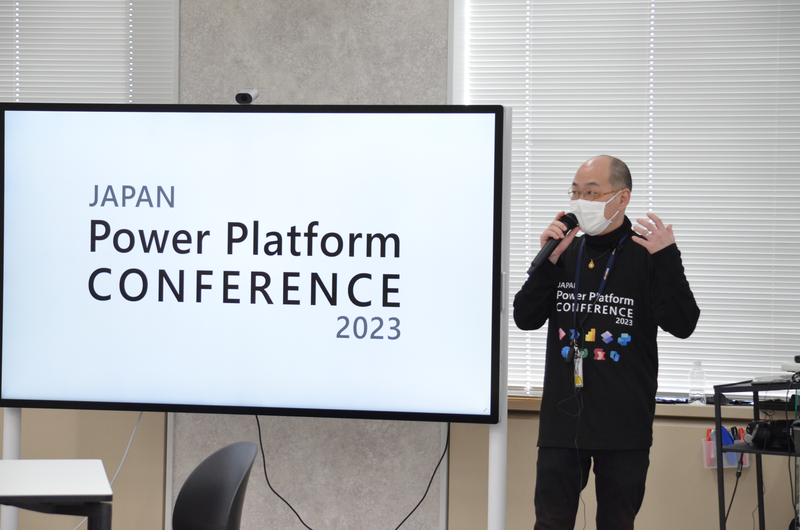 JAPAN Power Platform CONFERENCEで、男性の登壇者がマイクを持ちプレゼンテーションを行っている。