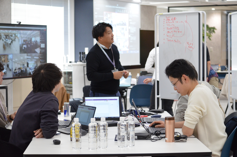 オフィスの会議スペースで複数の人がノートパソコンを使いながら議論している様子。オンライン会議の画面が映る大型モニターがあり、ホワイトボードには赤い文字でメモが書かれている。