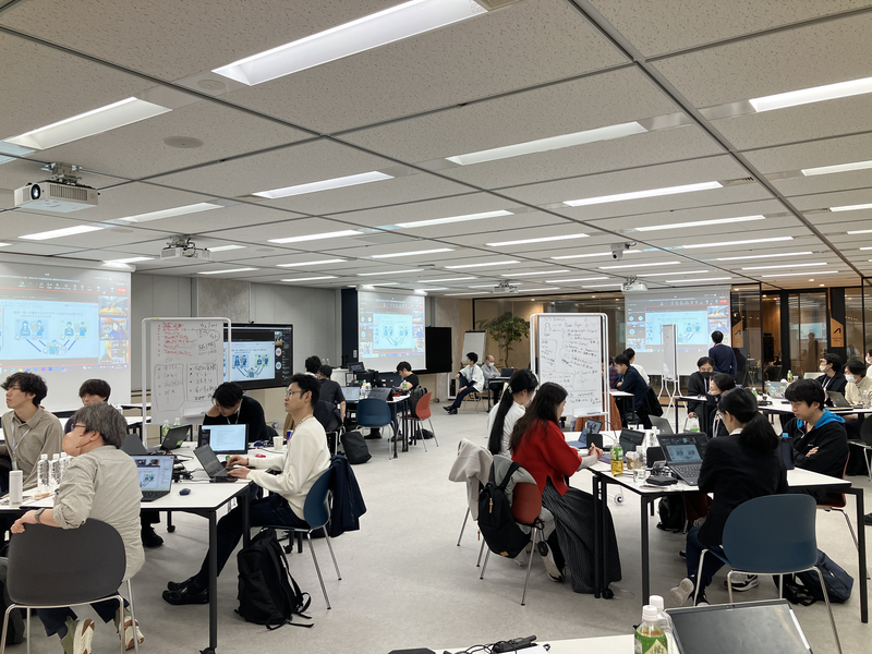 広いオフィススペースで複数のグループがテーブルに分かれてノートパソコンを使いながら作業や議論をしている様子。各テーブルには資料が置かれ、ホワイトボードには赤い文字でメモが書かれている。背景には複数の大型スクリーンにプレゼン資料が投影されている。
