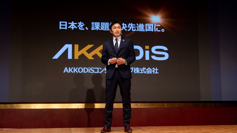 壇上に立つ男性。背後には「日本を、課題解決先進国に。AKKODiS」のスライドが映し出されている。