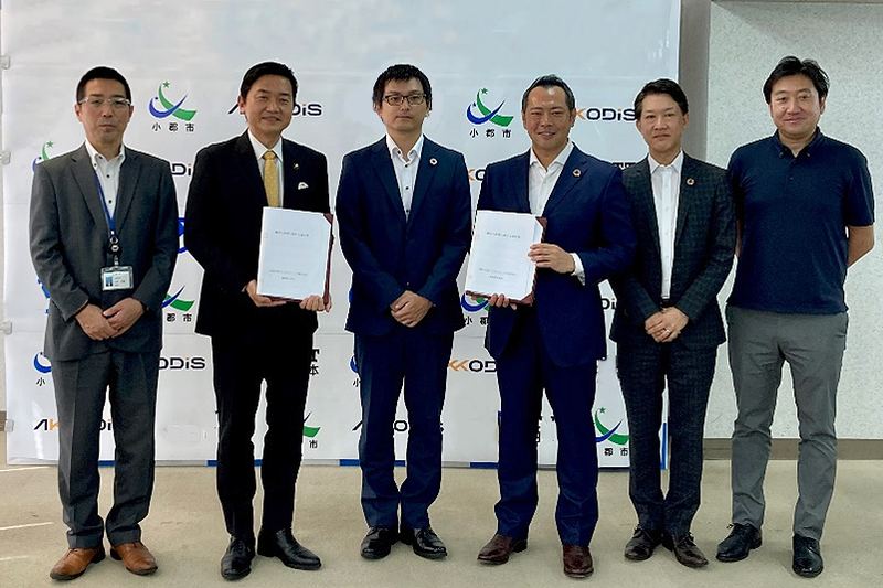 スーツを着た5人の男性と、右端にポロシャツを着た1人の男性が一列に並んでいる。左から二人目と、左から四人目の男性は、それぞれ書類を手に持っている。背後には小郡市のマークとAkkodisのロゴが入ったパネルが立てられている。